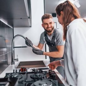 young-professional-plumber-in-grey-uniform-shows-results-of-work-for-housewife-on-the-kitchen-q25w1nopk1bplfeakirhyps008xh221aihxs0o6700